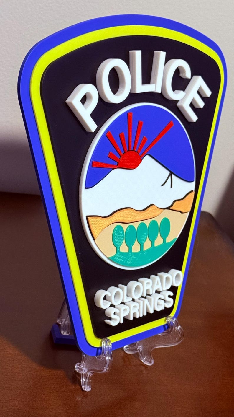 K&ouml;nnte beinhalten: Ein 3D-gedrucktes Polizeibadge mit blauem Hintergrund, einem wei&szlig;en Berg mit einer roten Sonne und gr&uuml;nen B&auml;umen. Das Abzeichen zeigt "POLICE" in wei&szlig;en Buchstaben und "COLORADO SPRINGS" in wei&szlig;en Buchstaben unten.