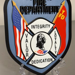 Op de afbeelding: Een 3D-geprint embleem voor de brandweer van Yonkers, met een blauwe en zwarte schildvorm. Het ontwerp bevat de tekst "YONKERS FIRE DEPARTMENT", vlaggen en de woorden "INTEGRITY", "VALOR", "DEDICATION" en "LOYALTY".