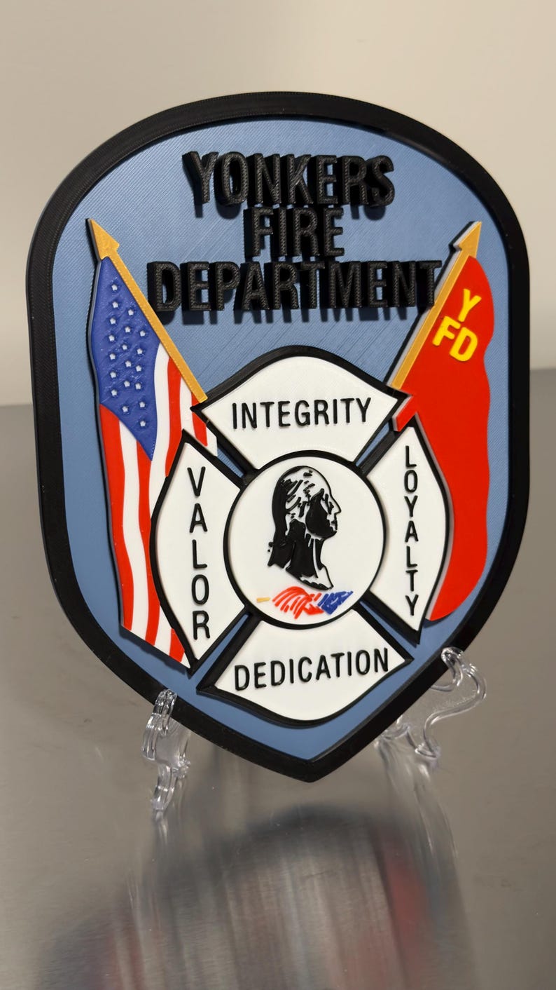 Op de afbeelding: Een 3D-geprint embleem voor de brandweer van Yonkers, met een blauwe en zwarte schildvorm. Het ontwerp bevat de tekst "YONKERS FIRE DEPARTMENT", vlaggen en de woorden "INTEGRITY", "VALOR", "DEDICATION" en "LOYALTY".