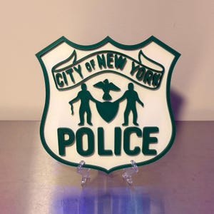 May include: A shield-shaped wall plaque featuring the City of New York Police emblem in dark green and off-white. The design includes the text CITY OF NEW YORK and POLICE, with two figures flanking a central shield and a bird icon above it.