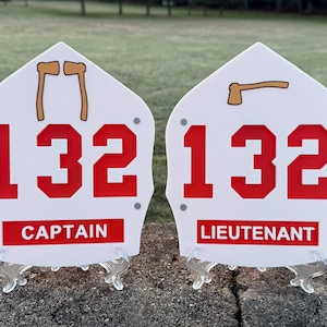 May include: Two white firefighter helmet plaques with red numbers 132. The plaque on the left says "Captain" and the plaque on the right says "Lieutenant".