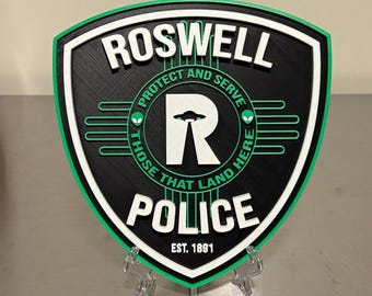 May include: A shield-shaped Roswell Police emblem in black, white, and green. The text reads ROSWELL POLICE, EST. 1891, and PROTECT AND SERVE THOSE THAT LAND HERE. A large white letter R features a small black UFO silhouette in the center.