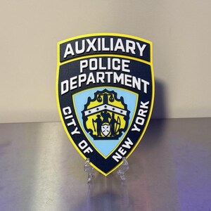 May include: A shield-shaped plaque featuring the Auxiliary Police Department City of New York emblem. The design includes bold white text on a dark blue background, a central yellow crest with black figures, and a bright yellow border trim.