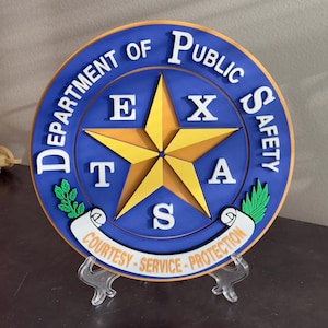 May include: A blue and gold circular sign with the words "Department of Public Safety" and "Texas" in white lettering. The sign features a five-pointed star with a scroll at the bottom that reads "Courtesy - Service - Protection".