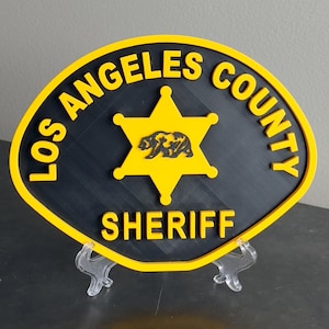 May include: A yellow and black Los Angeles County Sheriff's badge with a five-pointed star and a bear silhouette. The badge is on a clear plastic stand.
