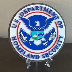 May include: A white round plaque with a blue eagle and the text "U.S. Department of Homeland Security" in a red and blue circle. The plaque is on a clear plastic stand.