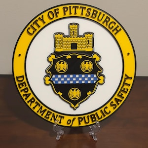 May include: A round, yellow and white sign with the text "City of Pittsburgh Department of Public Safety". The sign features a black and white crest with a castle, a checkered pattern, and three eagles.