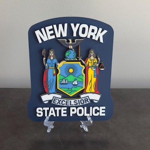 May include: A blue and white 3D wooden plaque with the words "NEW YORK STATE POLICE" and the New York State seal. The seal features a shield with a sun, a ship, and two women holding a staff and a scale of justice.