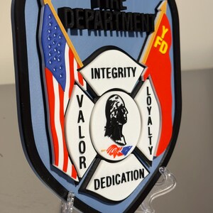 Op de afbeelding: Een schildvormig embleem voor de brandweer van Yonkers, met de Amerikaanse vlag en een rode vlag met "Y FD". Het embleem bevat de woorden "Integriteit", "Moed", "Loyaliteit" en "Toewijding".