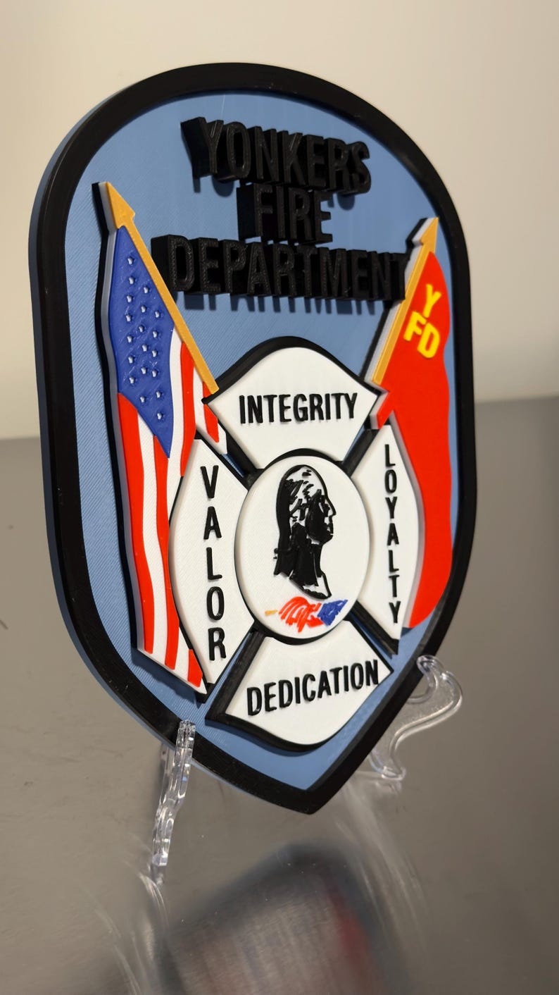 Op de afbeelding: Een schildvormig embleem voor de brandweer van Yonkers, met de Amerikaanse vlag en een rode vlag met "Y FD". Het embleem bevat de woorden "Integriteit", "Moed", "Loyaliteit" en "Toewijding".