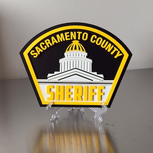 May include: A Sacramento County Sheriff badge with a black background and yellow border. The badge features the words "Sacramento County" in a yellow arc, and "SHERIFF" in yellow. A white building is in the center.