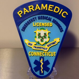 Op de afbeelding: Een blauw en geel Paramedic-badge met de tekst "Emergency Medical Services Licensed" en "Connecticut". De badge heeft een staatszegel en een levensster-symbool. De badge wordt op een transparante standaard weergegeven.