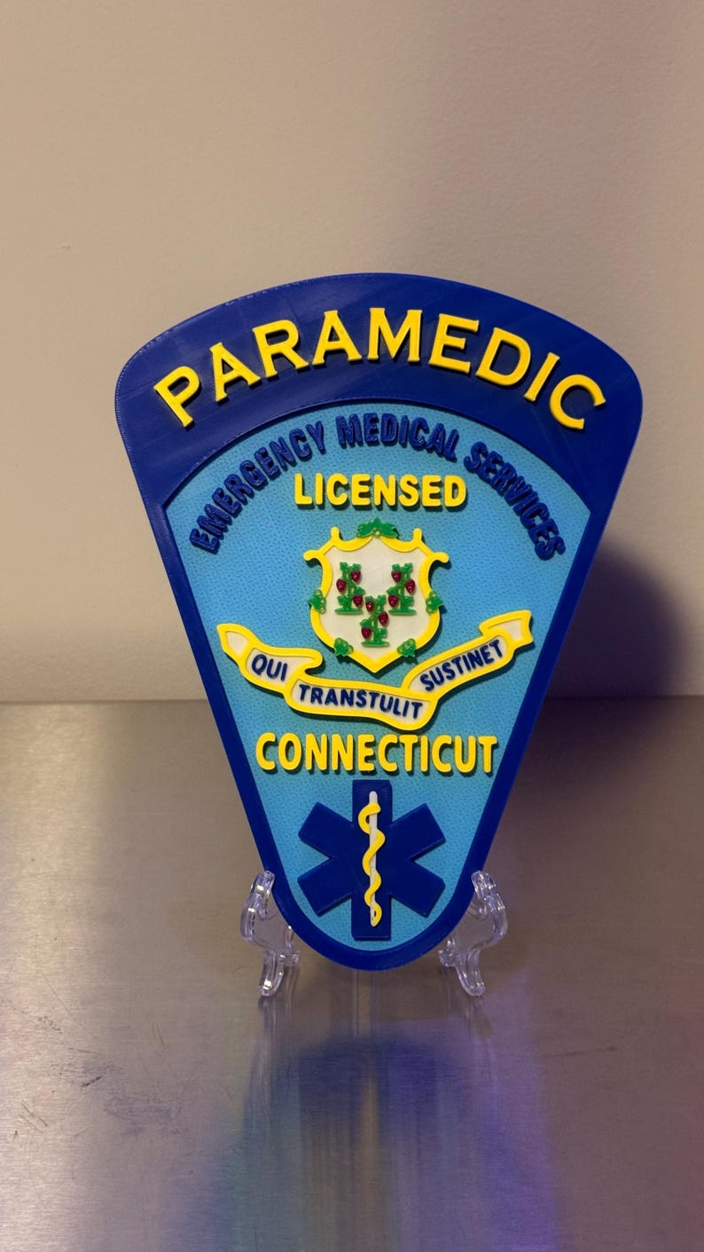 Op de afbeelding: Een blauw en geel Paramedic-badge met de tekst "Emergency Medical Services Licensed" en "Connecticut". De badge heeft een staatszegel en een levensster-symbool. De badge wordt op een transparante standaard weergegeven.