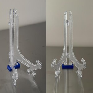 Op de afbeelding: Transparante acryl display stands met een decoratief scroll ontwerp. De stands hebben een blauwe basis en zijn ontworpen om borden of andere items vast te houden. De stands worden vanuit twee verschillende hoeken getoond.