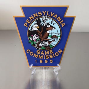 May include: A blue and gold Pennsylvania Game Commission sign. The sign features a deer, eagle, and turkey within a circular emblem. The words "Pennsylvania Game Commission 1895" are displayed on the sign.