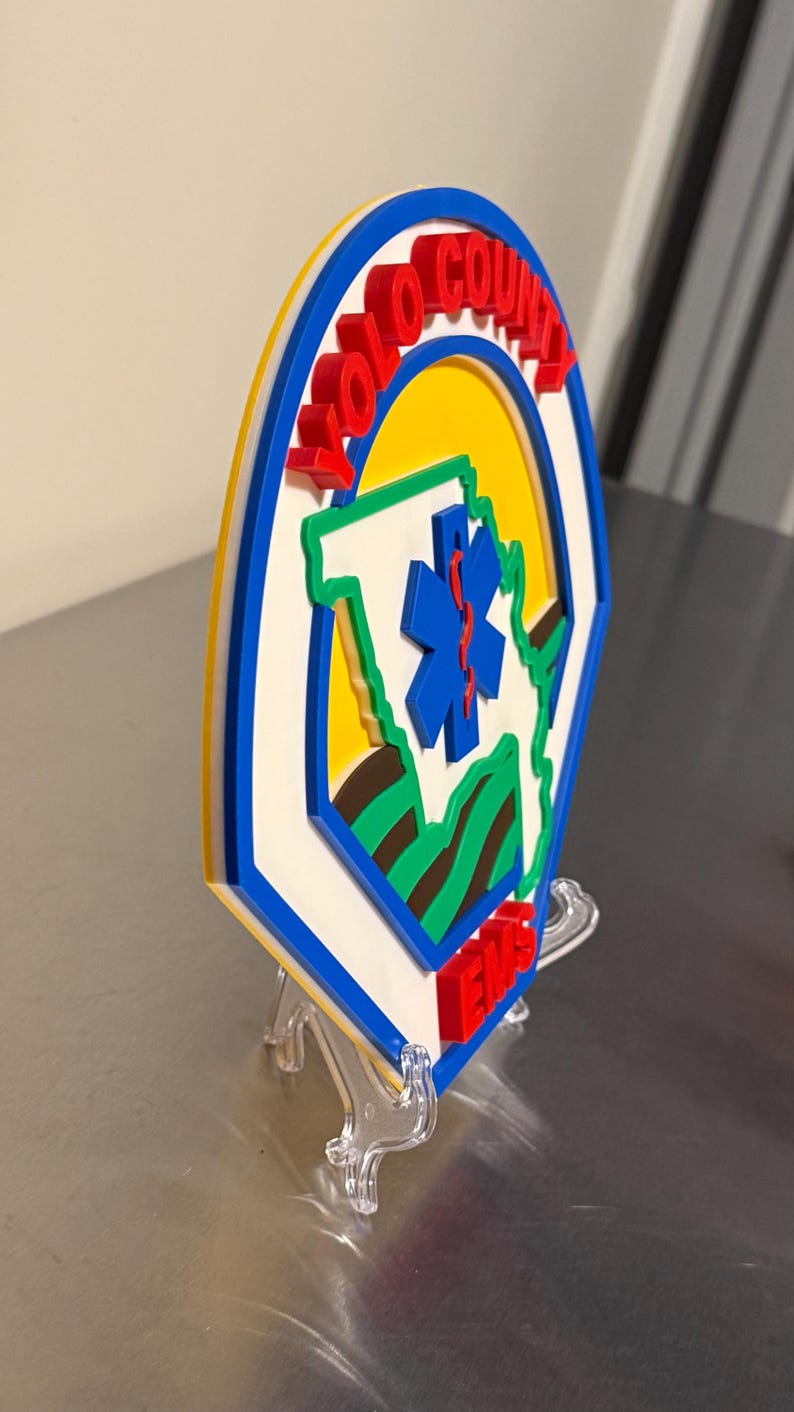 May include: A layered emblem with the text "YOLO COUNTY" in red, and "EMS" in red at the bottom. The design includes a blue star of life, a green outline of a state, and a yellow background, within a blue and white border.