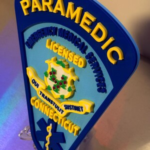 Op de afbeelding: Een blauw en geel Paramedic-bord met de tekst "PARAMEDIC EMERGENCY MEDICAL SERVICES LICENSED CONNECTICUT" en een symbool van de levensster. Het bord heeft een 3D-effect en is op een transparante standaard gemonteerd.
