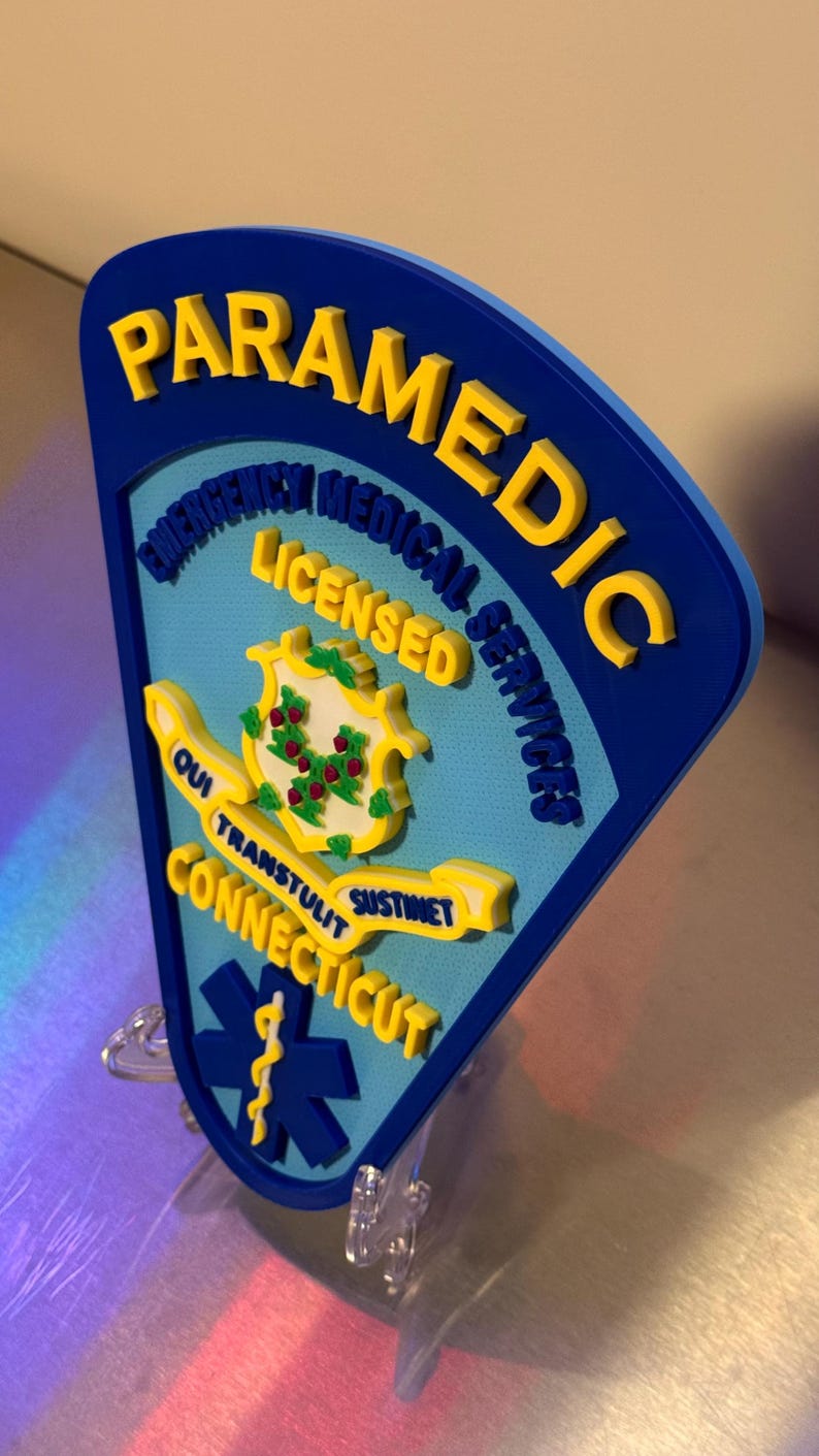Op de afbeelding: Een blauw en geel Paramedic-bord met de tekst "PARAMEDIC EMERGENCY MEDICAL SERVICES LICENSED CONNECTICUT" en een symbool van de levensster. Het bord heeft een 3D-effect en is op een transparante standaard gemonteerd.