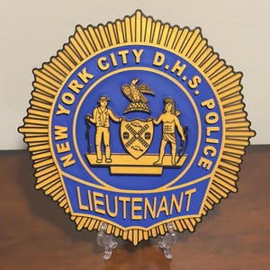 May include: A gold and blue New York City D.H.S. Police badge with a starburst design. The badge features a coat of arms with a shield and two figures. The text "LIEUTENANT" is printed on the badge.