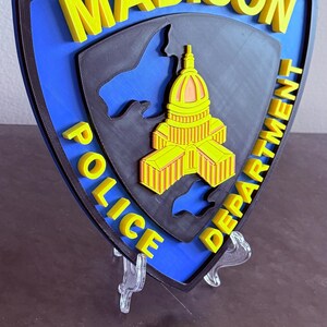 May include: A 3D-printed police badge with the words "MADISON POLICE DEPARTMENT" in yellow. The badge has a blue background with a black outline and a detailed yellow and orange building design in the centre.