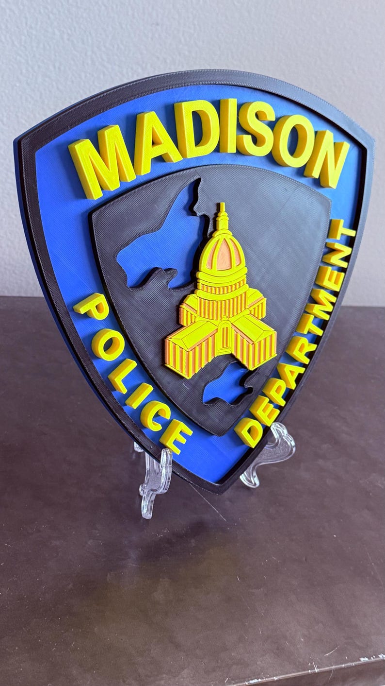 May include: A 3D-printed police badge with the words "MADISON POLICE DEPARTMENT" in yellow. The badge has a blue background with a black outline and a detailed yellow and orange building design in the centre.