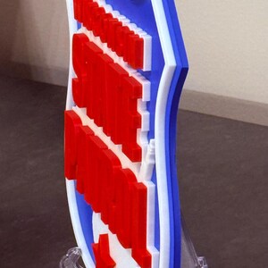 May include: A 3D-printed shield-shaped emblem in red, white, and blue. The design features a red star and text on a white background, framed by a blue border with white accents. It is displayed on a clear stand.