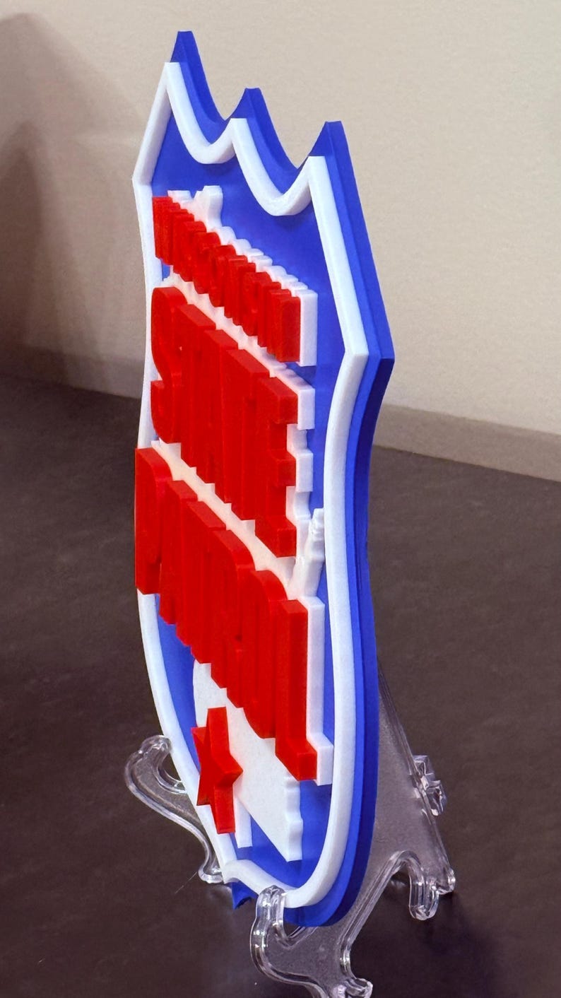 May include: A 3D-printed shield-shaped emblem in red, white, and blue. The design features a red star and text on a white background, framed by a blue border with white accents. It is displayed on a clear stand.