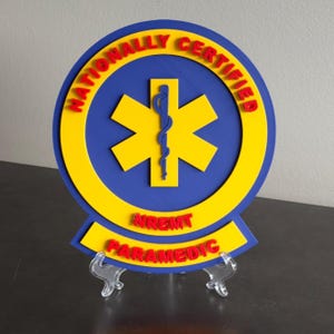 May include: A circular sign featuring a blue background with a yellow Star of Life emblem and a central blue Rod of Asclepius. The text NATIONALLY CERTIFIED is in red along the top, with NREMT and PARAMEDIC in red on a yellow banner at the base.