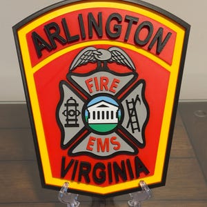 May include: A red and yellow 3D printed patch with the words "ARLINGTON FIRE EMS VIRGINIA" and a stylized eagle with wings spread above a cross with a fire hydrant, building, and ladder.