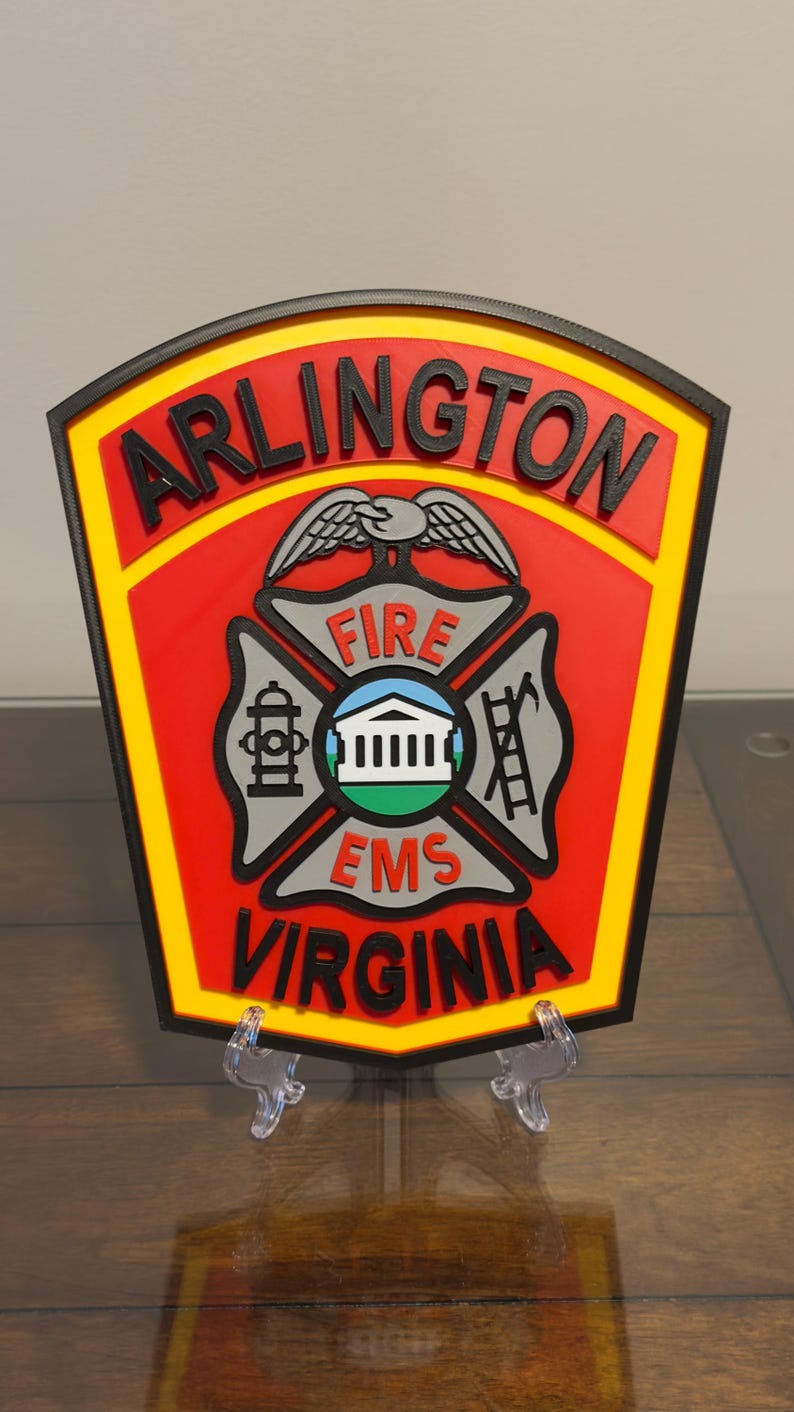 May include: A red and yellow 3D printed patch with the words "ARLINGTON FIRE EMS VIRGINIA" and a stylized eagle with wings spread above a cross with a fire hydrant, building, and ladder.