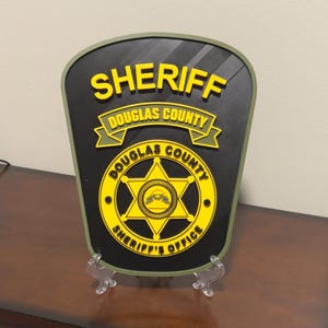 May include: A black and yellow sheriff's badge with the words "Sheriff Douglas County Sheriff's Office" on it. The badge is mounted on a green stand.