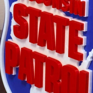 May include: A blue, white, and red sign featuring the words "Wisconsin State Patrol." The words are in bold, red letters, with a white outline. The sign has a white outline and a red star.