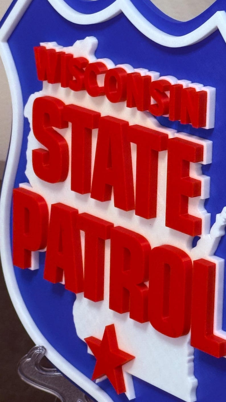 May include: A blue, white, and red sign featuring the words "Wisconsin State Patrol." The words are in bold, red letters, with a white outline. The sign has a white outline and a red star.