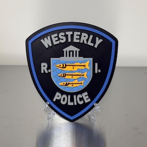 May include: A shield-shaped emblem with the words "Westerly R.I. POLICE" in white lettering. The emblem features a blue border, a black background, and a central design of three yellow fish on a blue background.