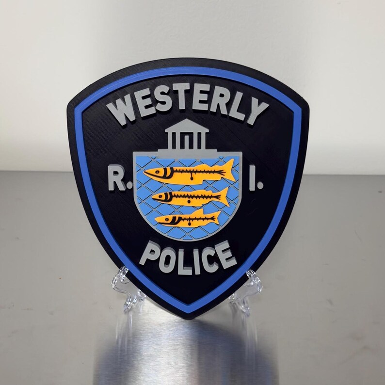 May include: A shield-shaped emblem with the words "Westerly R.I. POLICE" in white lettering. The emblem features a blue border, a black background, and a central design of three yellow fish on a blue background.