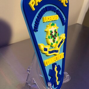 Op de afbeelding: Een blauw en geel paramedicus embleem met de woorden "Paramedic", "Emergency Medical Services", "Licensed", "Qui Transtulit Sustinet" en "Connecticut". Het embleem heeft een levensster symbool en een staatszegel.