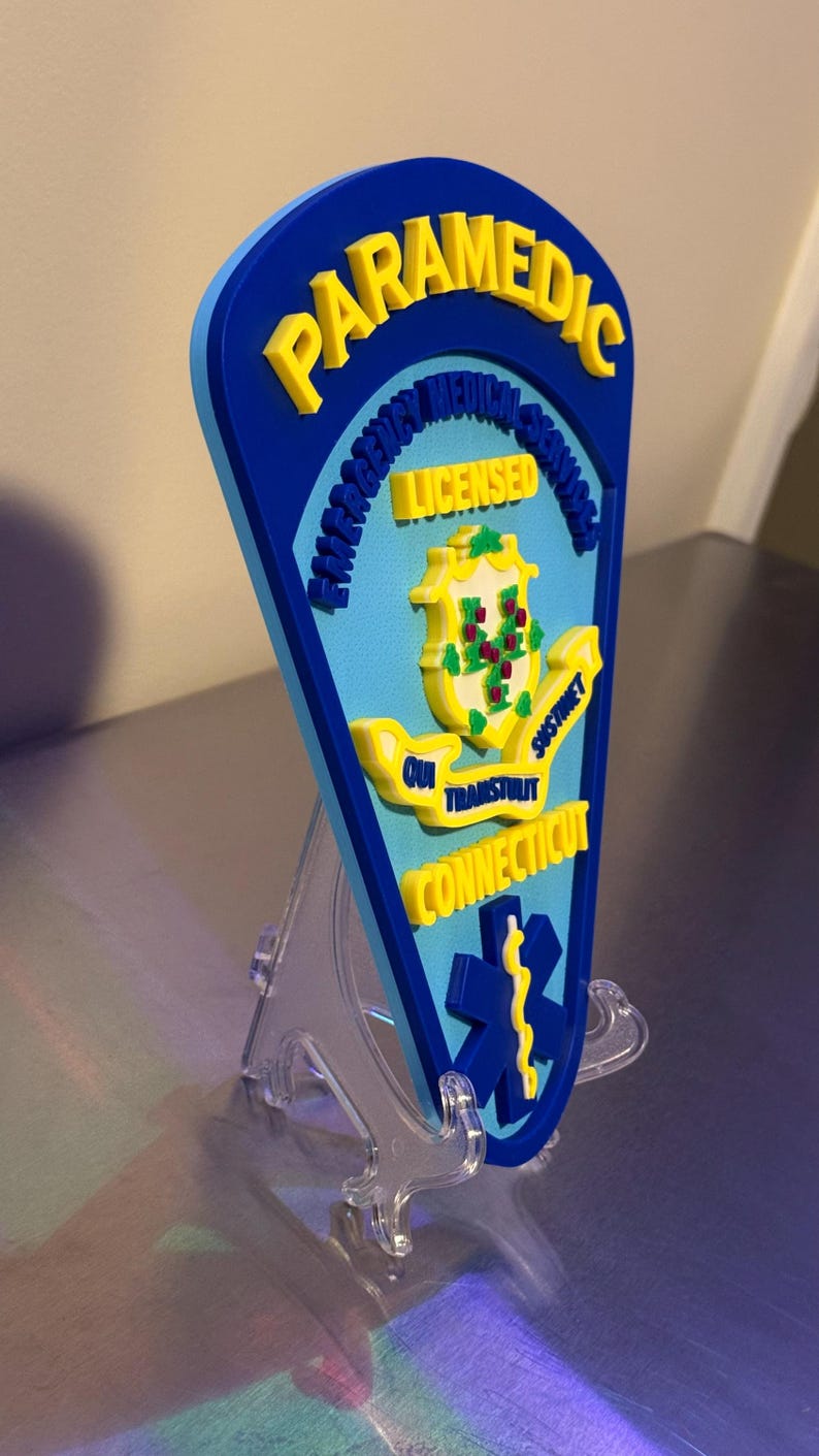 Op de afbeelding: Een blauw en geel paramedicus embleem met de woorden "Paramedic", "Emergency Medical Services", "Licensed", "Qui Transtulit Sustinet" en "Connecticut". Het embleem heeft een levensster symbool en een staatszegel.