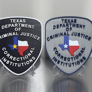May include: Two shield-shaped plaques featuring the Texas Department of Criminal Justice logo. One is black with white text and a colorful Texas map. The other is silver with dark blue text and a similar map. Both are displayed on clear stands.