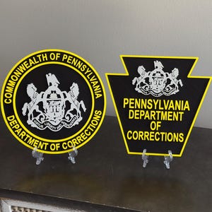 May include: Two black and yellow signs featuring the Commonwealth of Pennsylvania Department of Corrections emblem. The circular sign displays the state seal and text, while the keystone-shaped sign features the seal and department name.