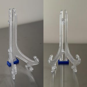 May include: Clear acrylic display stands with a decorative scroll design and blue bases. These stands are designed to hold plates, artwork, or other items. The stands are shown from the side and front.