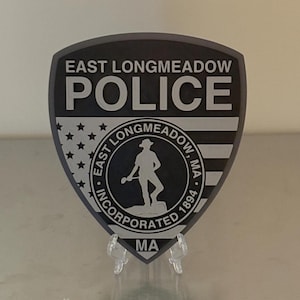 May include: A shield-shaped emblem for the East Longmeadow Police. The top reads "EAST LONGMEADOW POLICE" in white letters. Below is a design with stars and stripes, and a circular emblem with a figure and the text "EAST LONGMEADOW, MA INCORPORATED 1894".