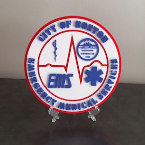 May include: A white round plaque with a red border and the text "City of Boston Emergency Medical Services" in a blue circle. The plaque features a blue medical symbol, a blue caduceus, and a blue heartbeat graphic. The plaque is on a clear plastic stand.