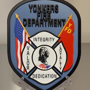 Op de afbeelding: Een schildvormig embleem voor de brandweer van Yonkers. Het embleem toont bovenaan de tekst "YONKERS FIRE DEPARTMENT", met een Amerikaanse vlag en een rode vlag met "FD" aan weerszijden. Het midden heeft een Maltezer kruis met de woorden "INTEGRITY", "VALOR", "DEDICATION" en "LOYALTY".