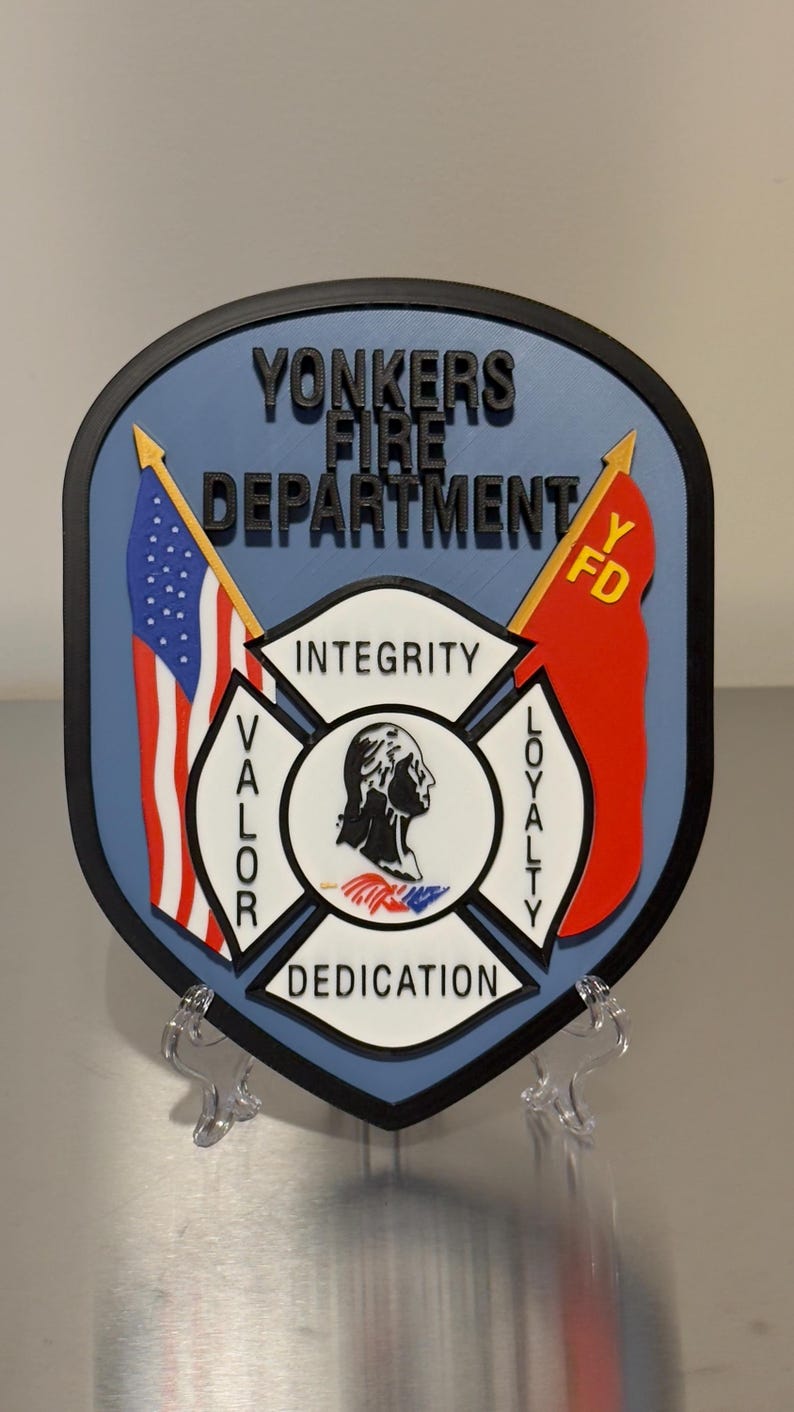 Op de afbeelding: Een schildvormig embleem voor de brandweer van Yonkers. Het embleem toont bovenaan de tekst "YONKERS FIRE DEPARTMENT", met een Amerikaanse vlag en een rode vlag met "FD" aan weerszijden. Het midden heeft een Maltezer kruis met de woorden "INTEGRITY", "VALOR", "DEDICATION" en "LOYALTY".