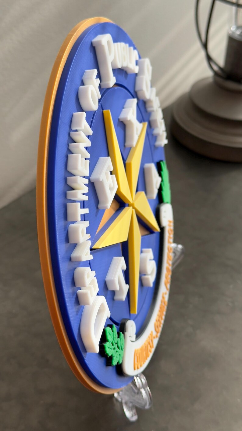 May include: A blue and yellow 3D printed wall plaque with the words "Department of Public Safety" and a star with the letters "E-9-1-1" around it. The plaque is on a clear plastic stand.