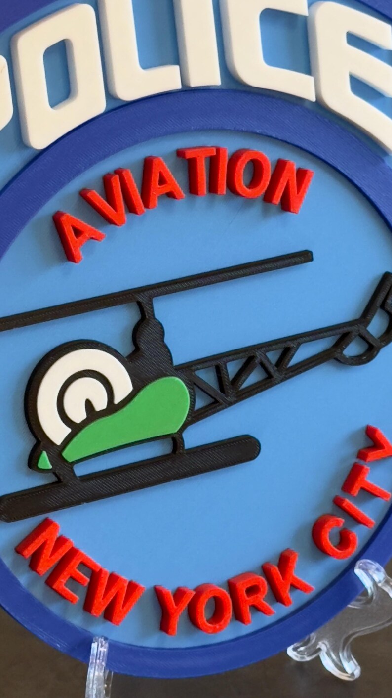 May include: A blue and white round sign with the words "POLICE AVIATION NEW YORK CITY" in red letters. A black helicopter with a green and white rotor is in the center of the sign.