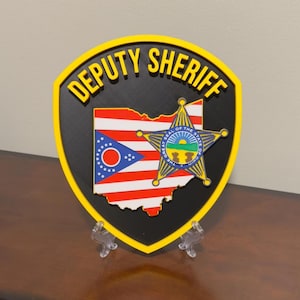 May include: A Deputy Sheriff badge-shaped plaque with a black background and yellow trim. The Ohio state flag is in the center, with a gold star and the words "Deputy Sheriff" in yellow.