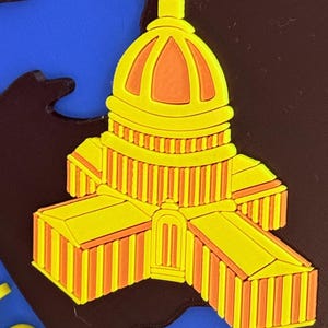 May include: A yellow and orange Capitol building design, possibly a magnet or sticker, set against a blue and brown backdrop. The building showcases detailed architectural features, including a dome and columns. The design is a vibrant representation of a landmark.