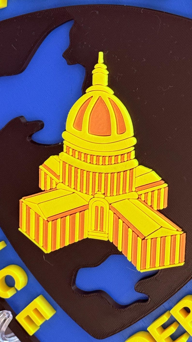 May include: A yellow and orange Capitol building design, possibly a magnet or sticker, set against a blue and brown backdrop. The building showcases detailed architectural features, including a dome and columns. The design is a vibrant representation of a landmark.