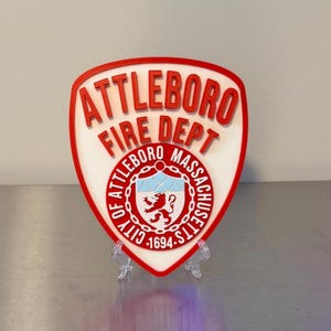 May include: A shield-shaped plaque featuring the red text ATTLEBORO FIRE DEPT above a circular emblem. The emblem displays a red lion, a chain border, and the text CITY OF ATTLEBORO MASSACHUSETTS 1694. The plaque has a white background and red border.